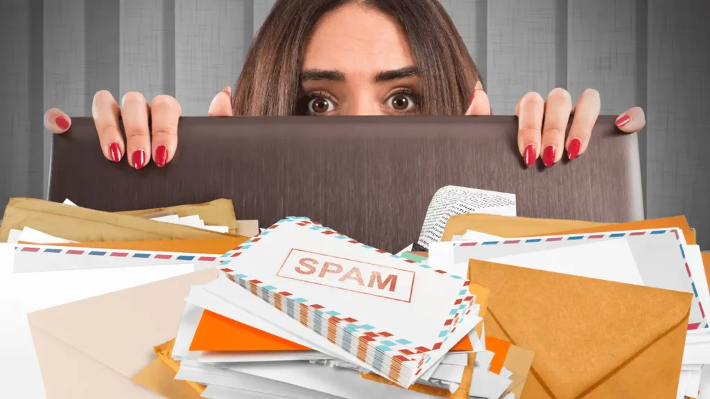 A woman with a scared expression hiding behind the back of a chair. In front of her are piles of envelopes, the middle pile says SPAM