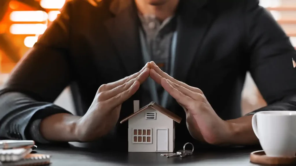 A person is sitting and on the table in front of them are notebooks, keys, a coffee cup, and a miniature model of a white house. The person is using their hands to mimic a roof over the house. 