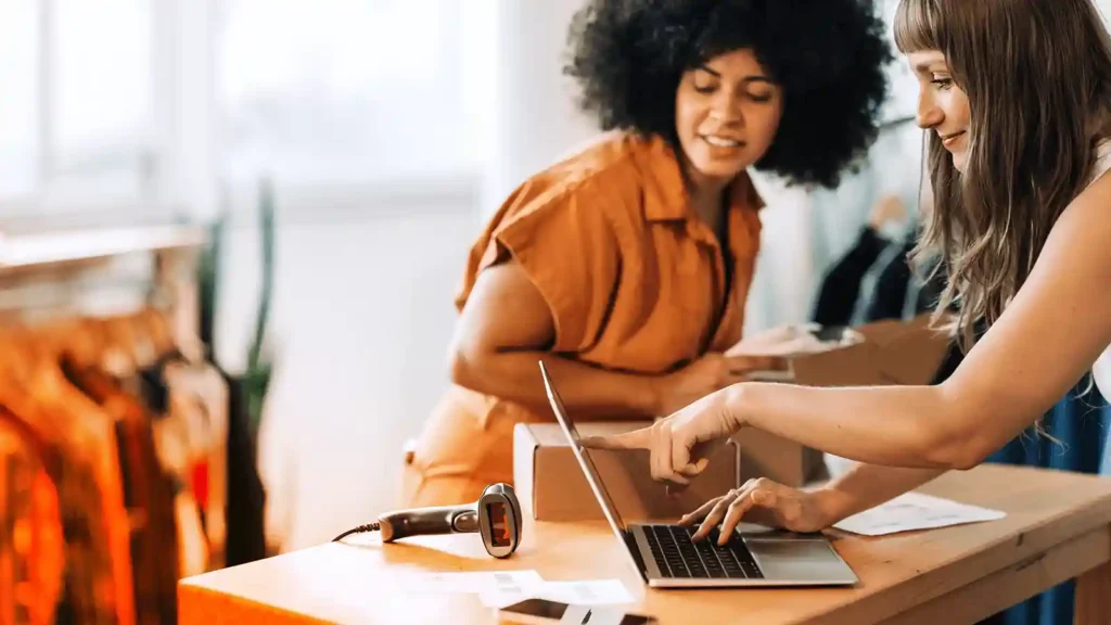 Two women working on a laptop while packing boxes and scanning labels. They appear to be in a clothing store.