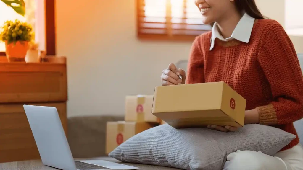 A woman sits on her couch with a box on her lap. She is writing out a label while looking at a laptop in front of her. Behind her are more packages and a dresser with a plant on top of it. 