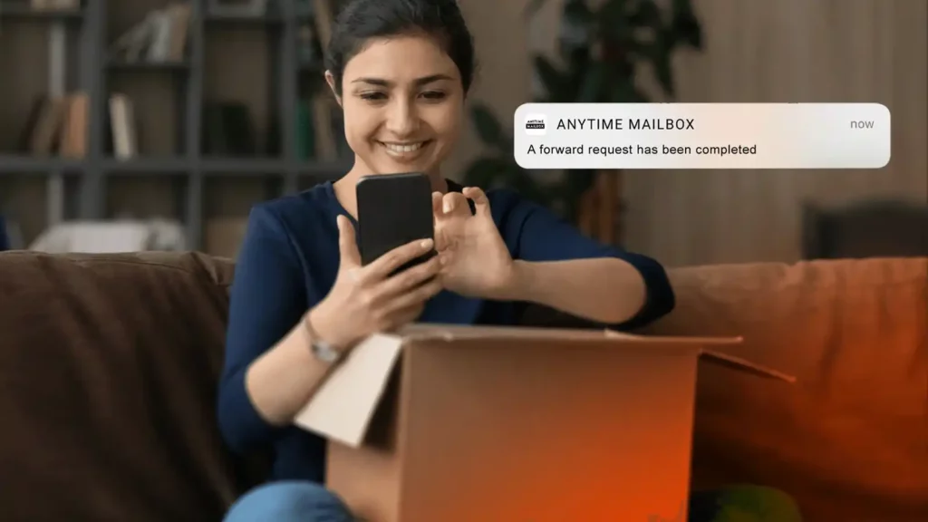 A woman with tan skin and hair pulled back, rests on a couch with a box on her lap, using her phone. Beside her is a picture of an Anytime Mailbox app notification with text that says “A forward request has been completed.” 