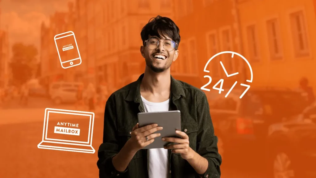 A man with dark hair smiles into the camera while using his tablet, beside him are icons that show a phone and laptop with the Anytime Mailbox logo and an icon that shows half a clock and 24/7