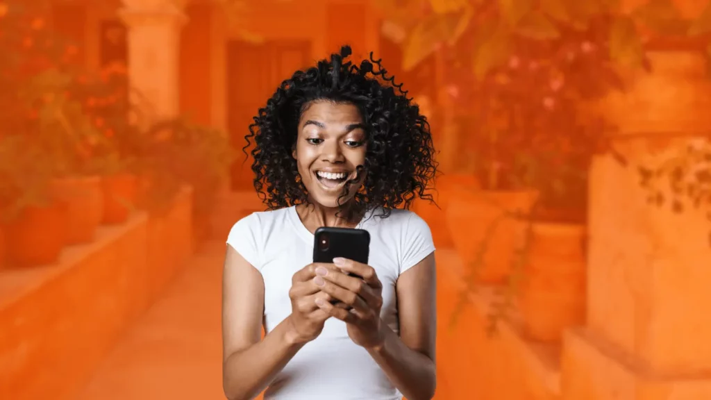 A woman with curly hair looks at her phone with an excited expression. Behind her is a stylized orange background of a garden path. 