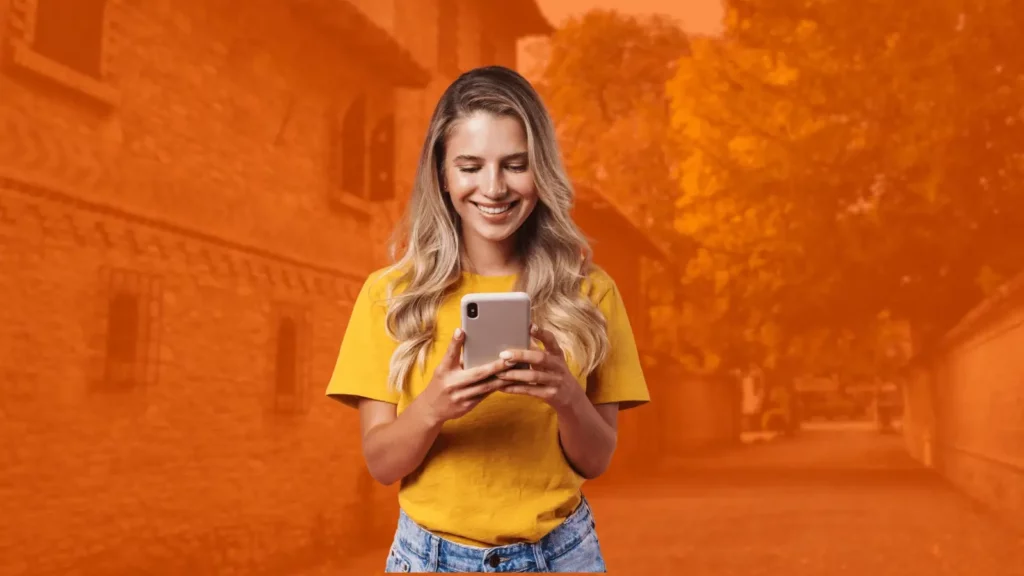 Woman in a yellow blouse using her phone, in the background is a stylized orange cobblestone street with brick houses and walls. 