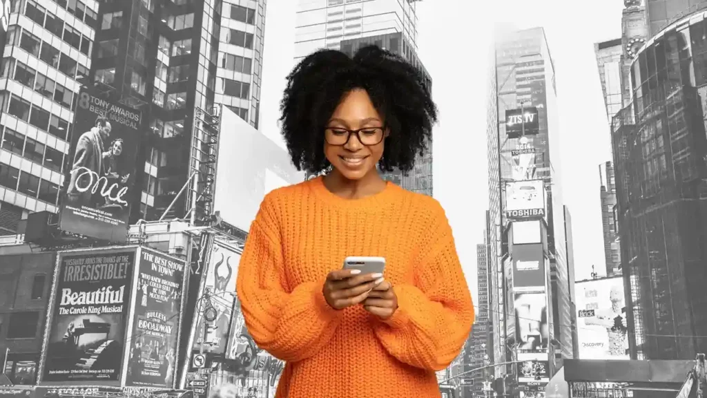 Woman in orange sweater using her phone, in the background is a black and white cityscape.