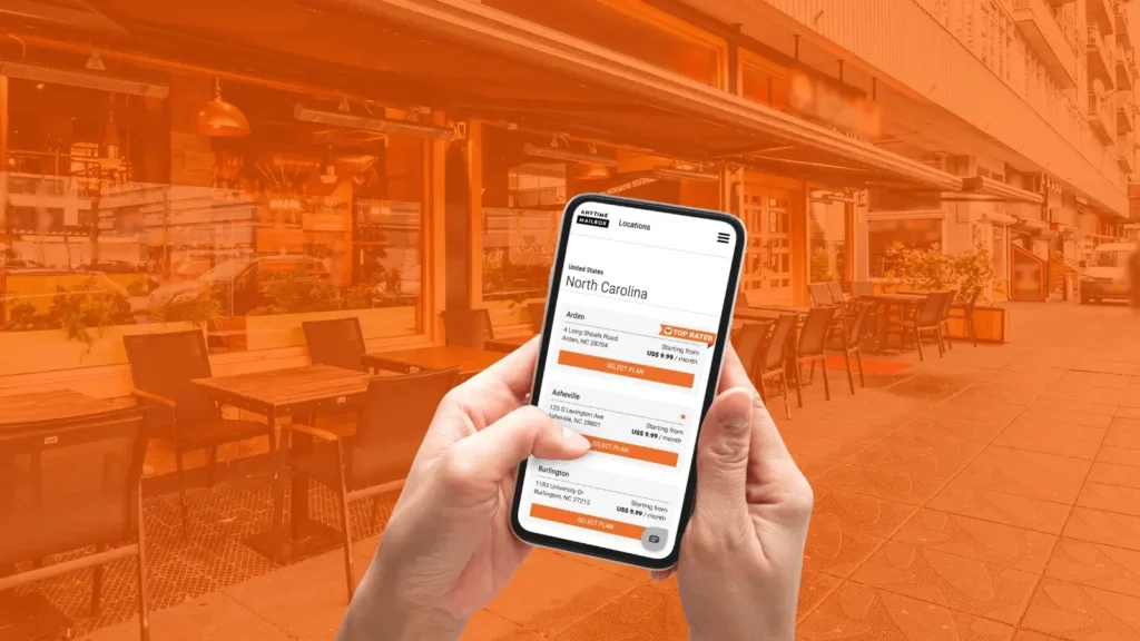 Close-up shot of a person’s hands using a phone while on a restaurant side street, with the Anytime Mailbox North Carolina webpage on screen. 