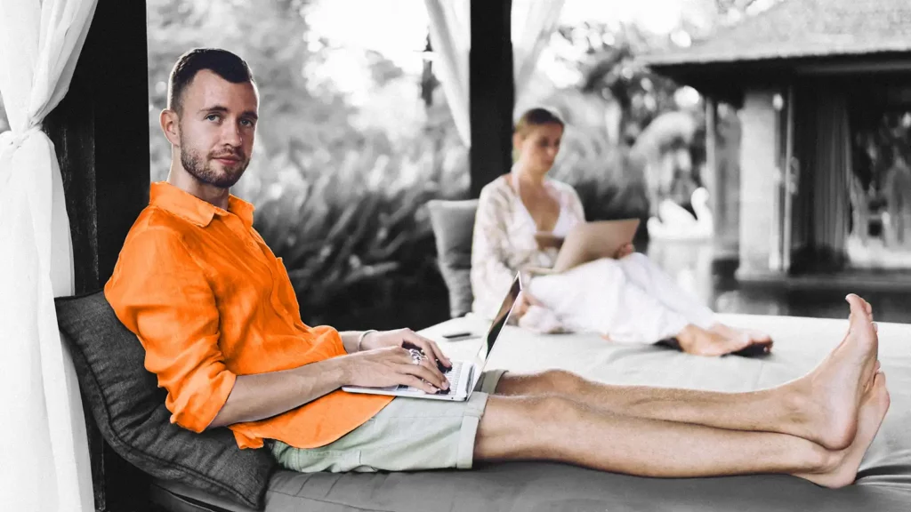 A man and woman lounging in a tropical setting. They are both using laptops.