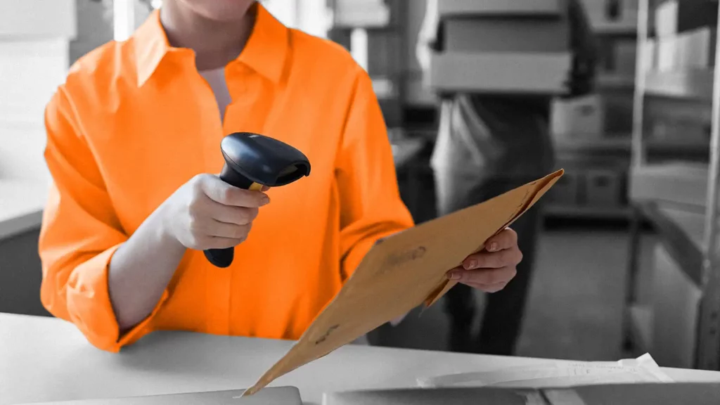 A person holding an envelop in one hand and a scanner in the other. Behind them are shelves of boxes.