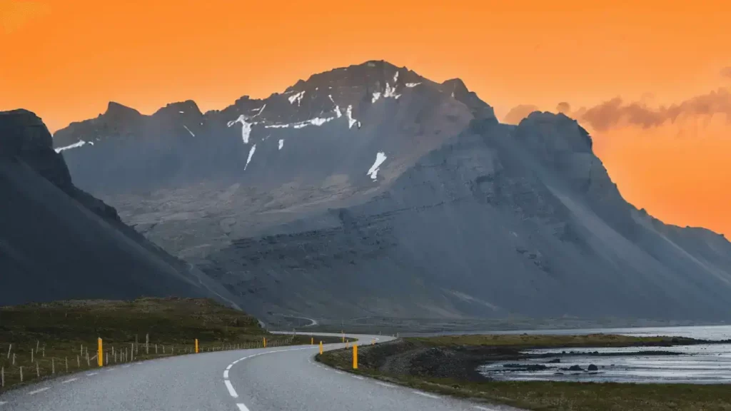 An empty winding highway beside a body of water and going towards a mountain range.