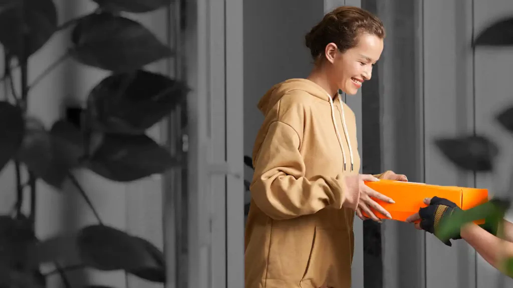 Woman in orange hoodie receiving an orange box. There are plants on the foreground of the image.