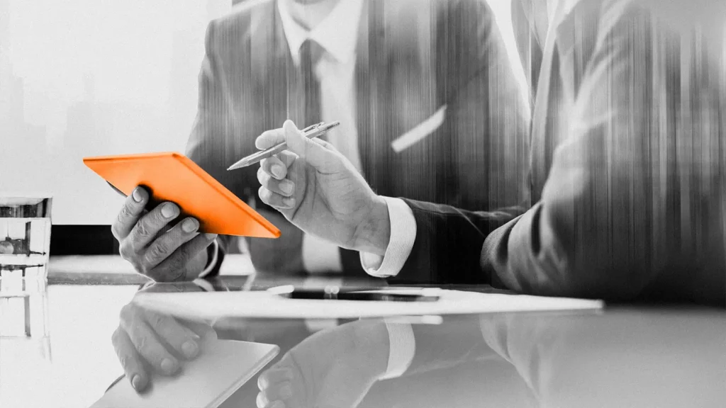 Two people engaged in a discussion, seated at work desks. The person on the left holds a stylized orange paper and the one on the right uses a pen to point to it. 