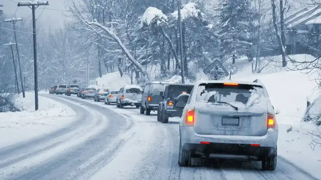 Line of cars traveling along a snowy road. The road is lined with trees and ground with snow.