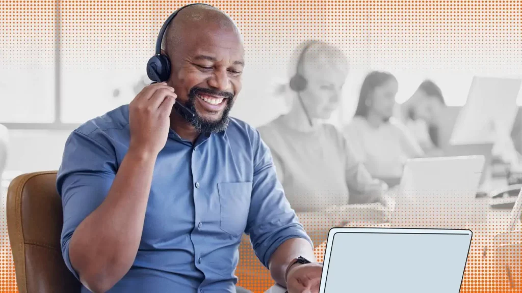 Smiling man wearing a headset and sitting in front of laptop. There are other people with headsets behind him.