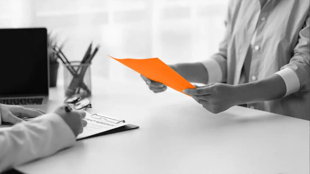 One pair of hands holding an orange paper. Another pair writing on a clipboard.