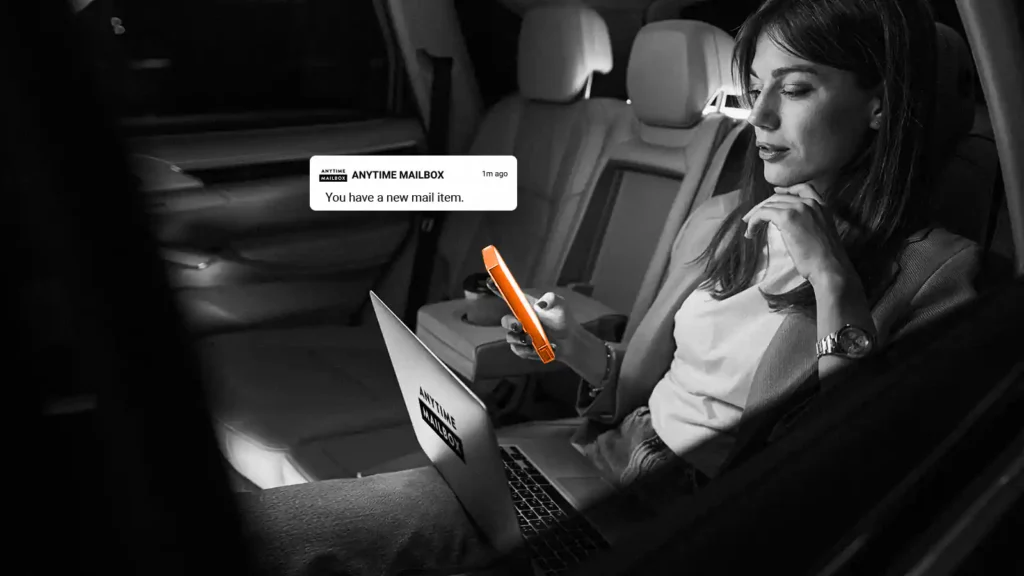 A woman in the backseat of a car. She has a laptop in front of her and is holding a mobile phone. There is an Anytime Mailbox pop-up near the phone.