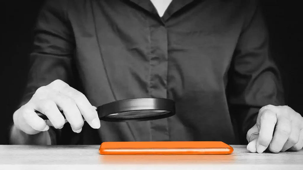 Man holding a magnifying glass over the screen of a mobile phone that’s on a flat surface.