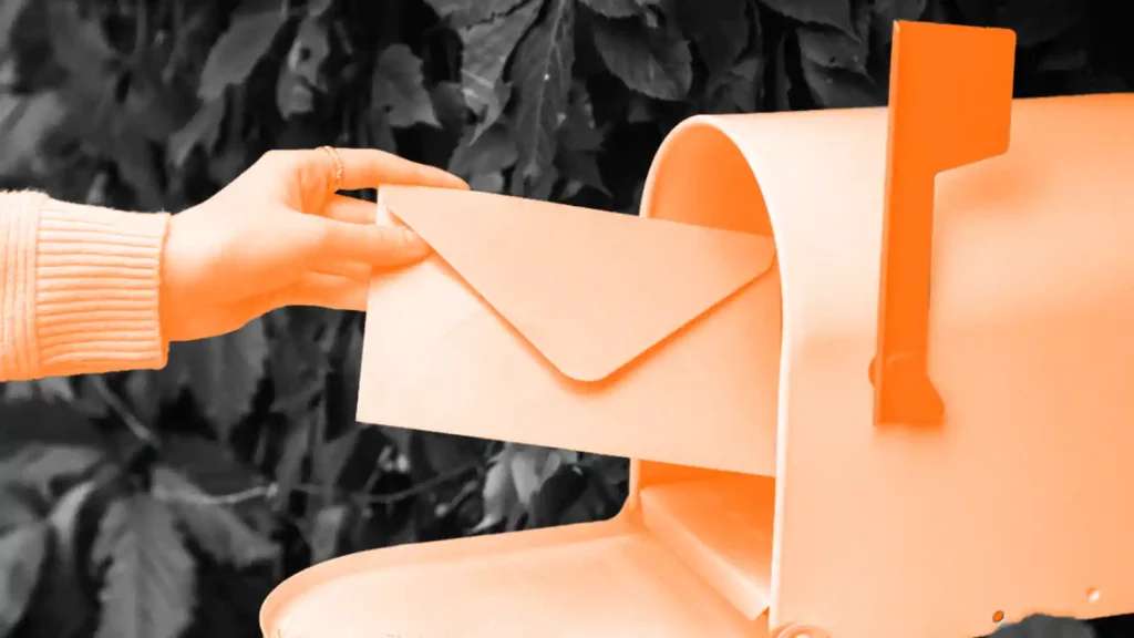 Hand holding an envelope to an open orange mailbox door.